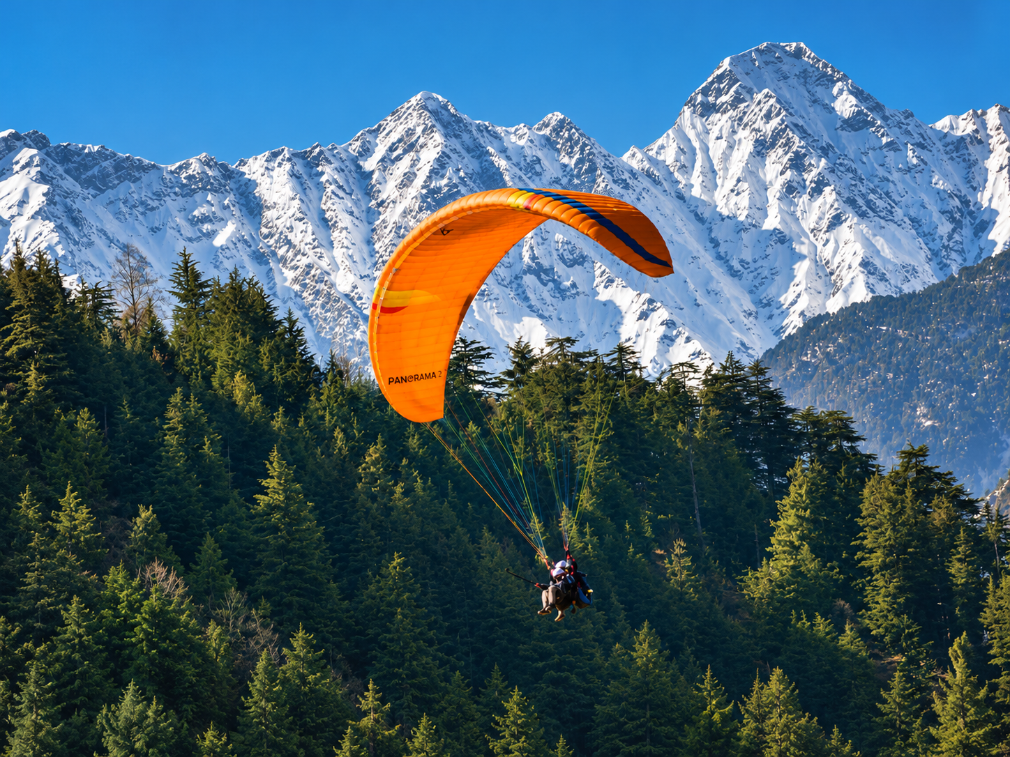 Orange tandem paraglider flying above pine forests with snow-covered mountains in Solang Valley.