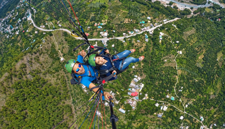 Overhead view of a tandem pilot and passenger flying above orchards, village roads and the river in Dobhi.